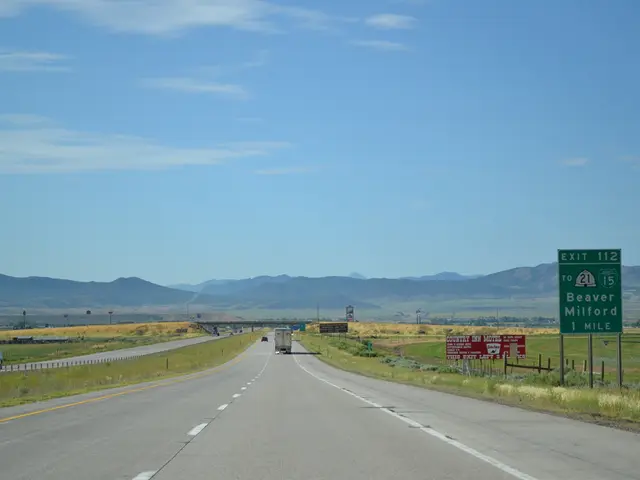 In this image we can see sky with clouds, hills, motor vehicles on the road, barrier poles, sign...