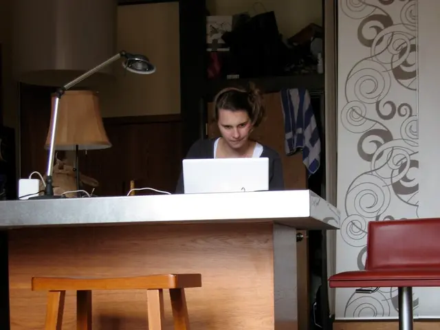 In this image there is a woman sitting in chair and operating a laptop , and on table there are...